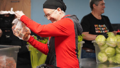 A smiling woman in a red sweater and black headscarf raising her arms indoors.