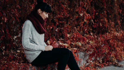 A woman in a gray sweater and burgundy scarf sits against a backdrop of vibrant red leaves.